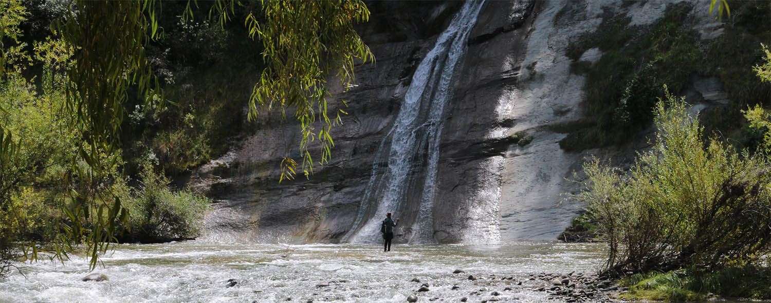 fly fisherman standing, dwarfed, in shallow water of river, below high waterfall