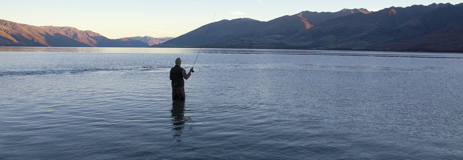 Fly fisherman at dusk, top of Lake Wanaka with mountains silhouetted against early evening sky